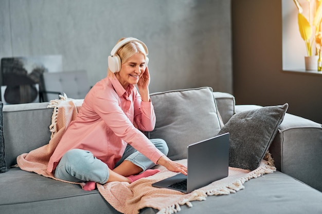 Video communication at a distance Beautiful senior woman uses laptop adjusts headphones and types on keyboard