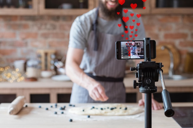 Video recipe Like feedback Virtual cooking training Blueberry pie Male chef blogger recording live streaming on smartphone on tripod in defocused loft kitchen with like hearts