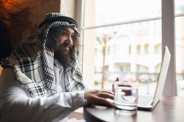 영상 통화. 사무실에서 일하는 아라비아 사업가, 장치, 가제트를 사용하는 비즈니스 센터. 현대 사우디 라이프 스타일. 전통 의상과 스카프를 입은 남자는 자신감 있고 바쁘고 잘 생겼습니다. 민족, 금융.