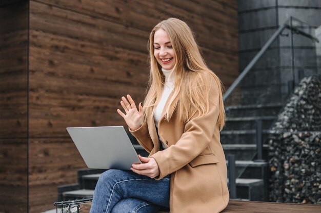 Videogesprek concept. jonge blanke blonde vrouw zittend op een bankje met een laptop voor een kantoor denemarken glimlachend en zwaaiend naar het beeldscherm