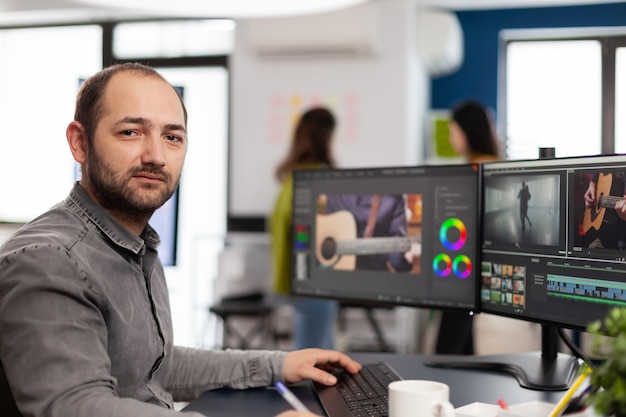 Videograaf die naar camera kijkt die glimlacht, werkt op een creatieve startup-werkplek met behulp van bewerkingssoftware voor postproductie op professionele pc met twee monitoren die videofilmmontage verwerken