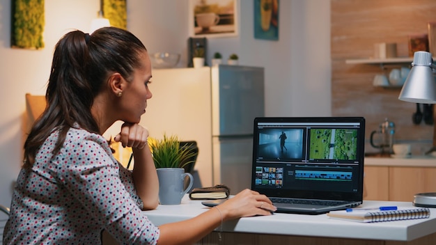 Videographer editing from home on professional laptop sitting on desk in modern kitchen in midnight. Creative video editor working at night at new project processing audio film montage.