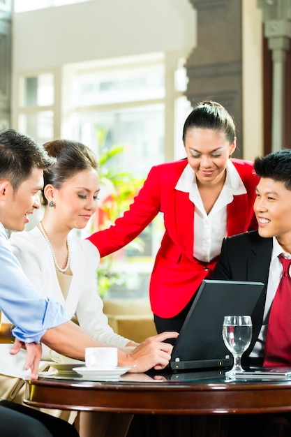 Vier aziatische chinese kantoormensen of zakenlieden en vrouwelijke ondernemers met een zakelijke bijeenkomst in de lobby van een hotel bespreken documenten op een tabletcomputer terwijl ze koffie drinken