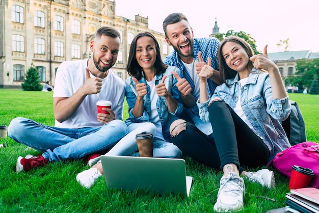 Vier beste jonge studentenvrienden met een laptop op het gras op universitair grondgebied zijn blij en opgewonden