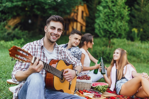 Vier jonge aantrekkelijke persoon chiiling in het park en een van hen speelt gitaar en smilling.