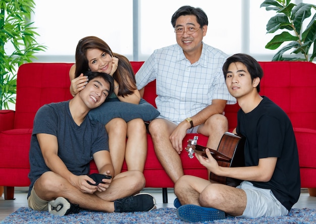 Vier leden Aziatische familie bestaande uit ouders, twee volwassen zonen die tijd doorbrengen met gitaar spelen, samen zingen met geluk op de bank tijdens vakanties in de woonkamer thuis. Ze kijken naar de camera.