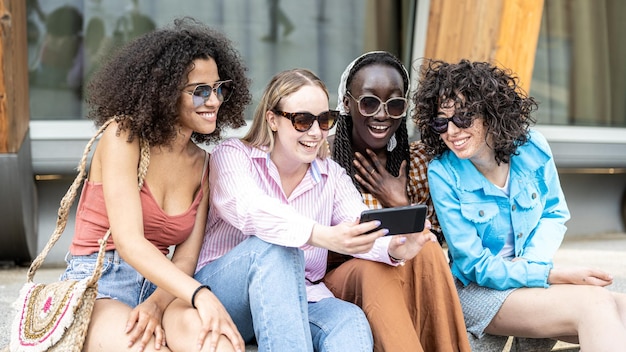 Foto vier meisjes, tieners van generatie z in de stad, jonge vrouwen die op een bankje in de stad zitten en een smartphone gebruiken voor een sociaal netwerk