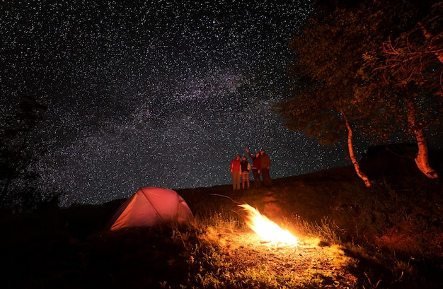 Vier personen genieten tijdens de nachtcamping van de ongewone hemel bezaaid met heldere sterren. Girl toont de rest van de groep op de Melkweg. Op de voorgrond brandt een vuur bij de tent en de bomen.