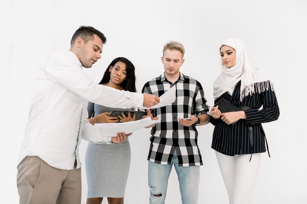 Vier vrienden van de Multiculturele Universiteit, mannelijke en vrouwelijke, staande op een witte achtergrond, terwijl blanke man papieren geeft om te studeren