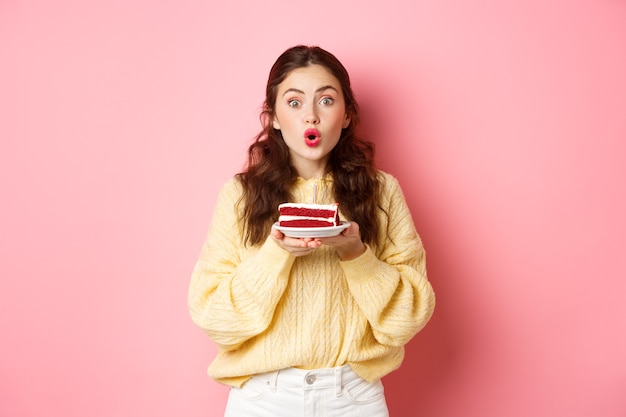 Viering en feestdagen. opgewonden feestvarken die b-day cake met kaars houden en verbaasd kijken naar de camera, staande tegen de roze muur.