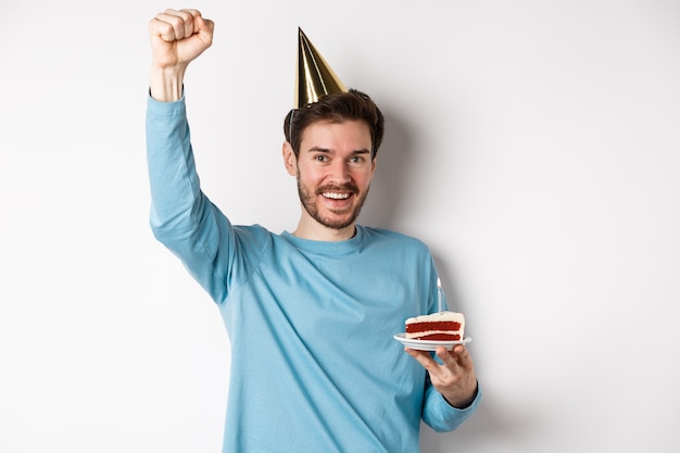 Viering en vakantie concept. Gelukkig man viert verjaardag in feestmuts, bday cake houden en hand opsteken in triomf, staande op witte achtergrond.