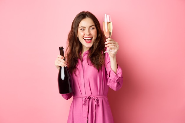 Viering en vakantie concept. Mooie vrouw die toast zegt, glas champagne opheft en fles vasthoudt, plezier heeft op feestje, staande tegen roze muur.