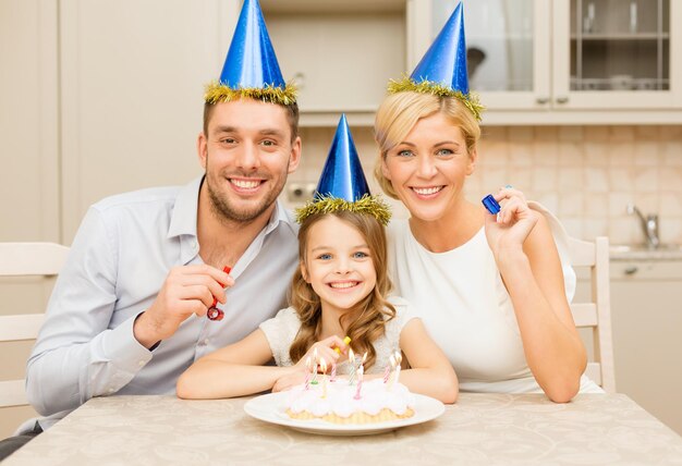 Viering, familie, vakantie en verjaardagsconcept - gelukkige familie in blauwe hoeden met cake en kaarsen die gunsthoorns blazen