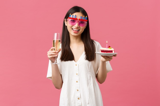 Viering, feestvakanties en leuk concept. Vrolijke gelukkige aziatische vrouw die verjaardag viert in grappige zonnebril, glas champagne en b-day cake met brandende kaars vasthoudt om een wens te doen.