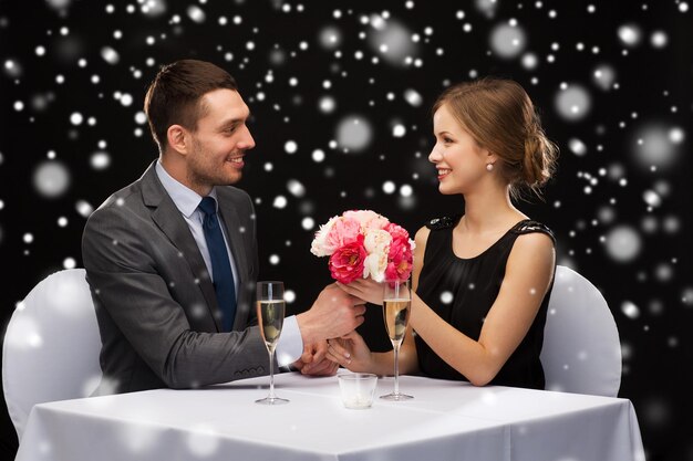 viering, kerstmis, vakantie en mensenconcept - glimlachend paar met champagne en bloemen bij restaurant over zwarte besneeuwde achtergrond