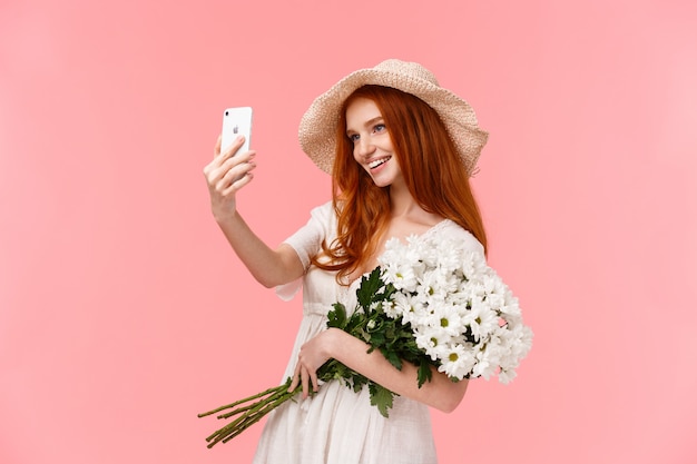 Viering, sociale media en internet concept. Verleidelijke brutale roodharige vrouw in strooien hoed, lente jurk, boeket houden, selfie nemen op smartphone met witte bloemen, tevreden glimlachen