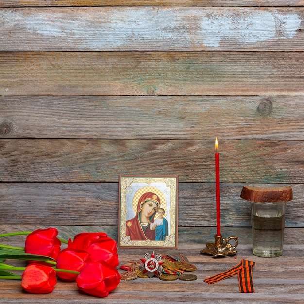 viering van overwinningsdagmedailles, orthodoxe icoon en brandende rode kaars, boeket bloemen van rode tulpen en een glas wodka met een stuk roggebrood