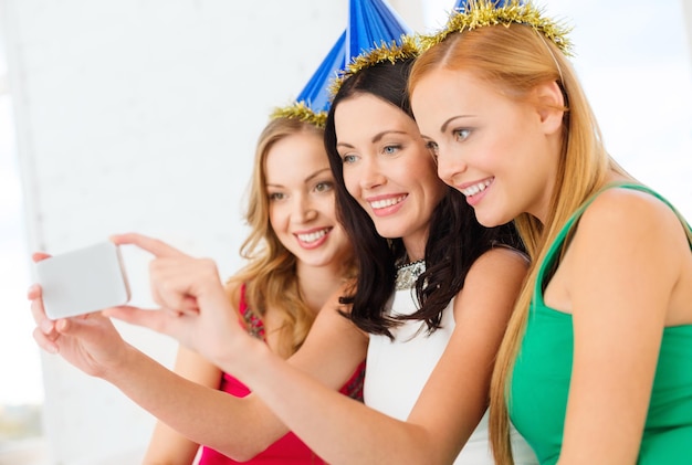 viering, vrienden, vrijgezellenfeest, verjaardagsconcept - drie lachende vrouwen in blauwe hoeden die plezier hebben met smartphone-fotocamera