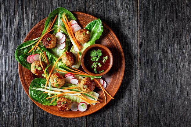 豚肉のミートボールにんじんきゅうりと大根のベトナムのバインミーサラダは、暗い素朴な木製の背景に甘いチリソースを添えた茶色のプレートのロマインレタスサラダに添えてください。