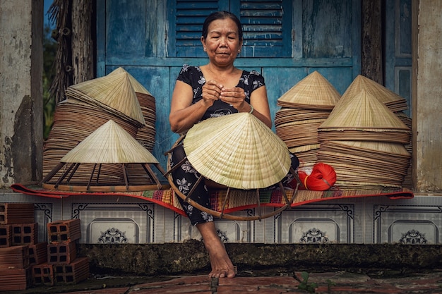 Вьетнамская старушка-ремесленница делает традиционную вьетнамскую шляпу в старом традиционном доме