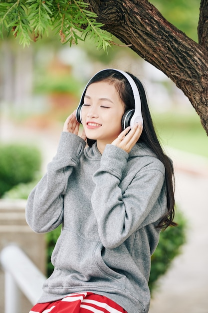 공원에서 나무 아래 앉아있을 때 헤드폰으로 음악을 듣고 즐기는 베트남 십 대 소녀