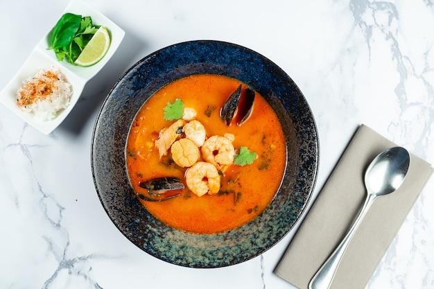 Вьетнамский суп Tom Yum с морепродуктами. Красный суп с креветками, морским гребешком, мидиями, лососем в темной стильной миске на мраморном столе.