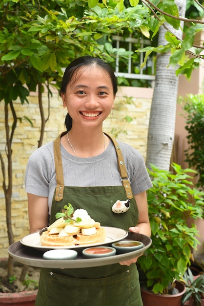 Vietnamese woman waitress serving Belgian waffles with banana and ice cream in outdoor cafe