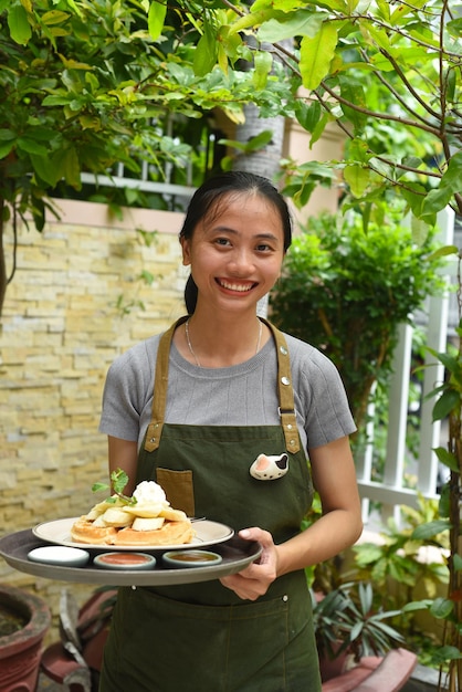 Vietnamese woman waitress serving Belgian waffles with fruits and ice cream in outdoor cafe