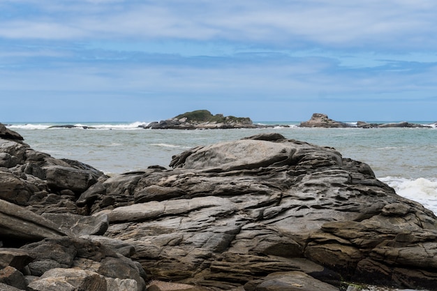 View of the beach of Rio das Ostras in Rio de Janeiro with sunny day, blue sky and some clouds. Strong sea and yellowish sand and lots of rocks.