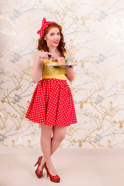 View of a beautiful pinup redhead girl holding a tray of colorful cupcakes.