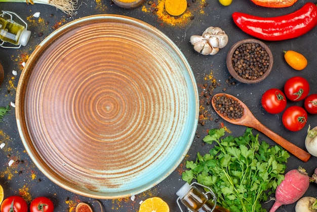 Above view of blue plate among fresh vegetables fallen oil bottle spices green bundle on dark color surface