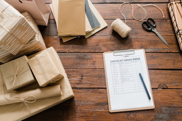 Photo view of checklist paper, pen, packed boxes, threads, scissors and stack of notepads on wood