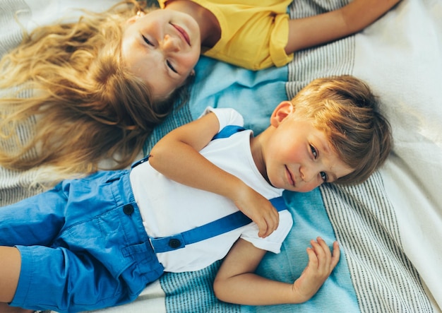 Above view of children lying on the blanket at the grass Happy little boy and cute girl enjoying summertime in the park Adorable kids smiling and having fun on sunlight outdoors Childhood