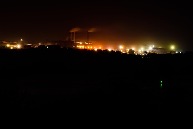 View on city lights at night. Smoke from chimneys of the factory rises to sky. Environmental pollution concept