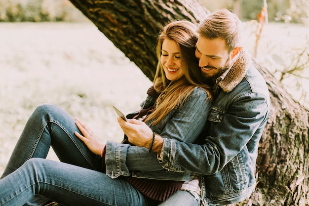 Вид на пара в любви, сидя на дереве в Осенний парк и с помощью мобильного телефона