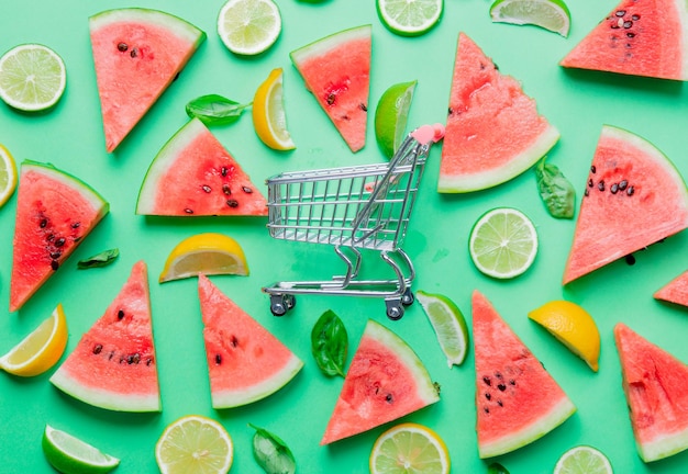 Above view at cutted lemons and limes with watermelon near shopping cart on green background