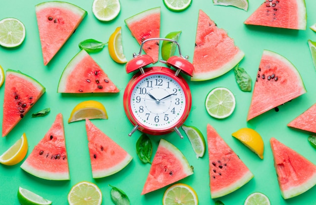 Above view at cutted lemons and limes with watermelon with alarm clock on green background