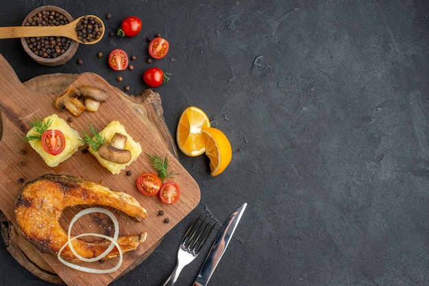 Above view of delicious fried fish and mushrooms tomatoes greens on cutting board cutlery set pepper on black surface