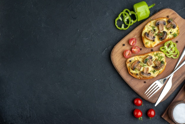 Above view of delicious snacks with mushroom fresh vegetables and cutlery set on wooden cutting board spices on the left side on black background