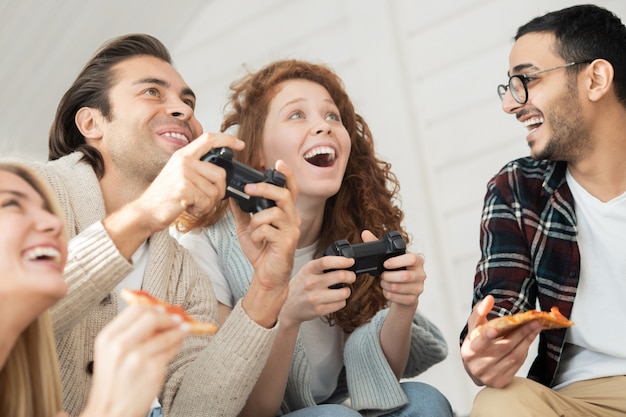 Below view of excited young man and woman playing video game while their friends eating pizza and rooting for them