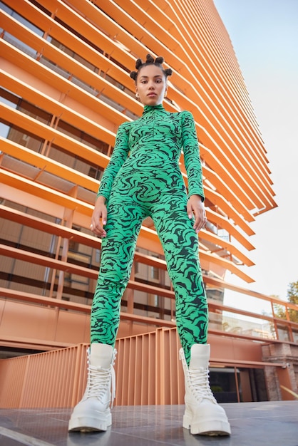 Below view of fashionable teenage girl dressed in green jumpsuit wears white shoes has trendy hairstyle poses against modern building looks confident at camera. Young people and fashion concept