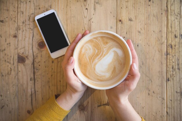 Sopra la vista delle mani femminili che tengono una tazza di caffè calda e con uno smartphone con un tavolo di legno