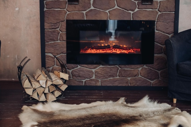 View over fireplace with burning logs, natural fur skin on the floor next to holder with logs in cozy room.