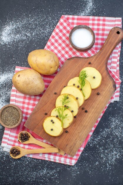 Above view of fresh chopped potatoes with dill pepper on wooden cutting board on red stripped towel spices on dark color background
