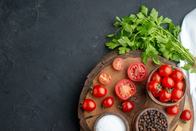 Above view of fresh tomatoes and spices in bowls spoons on wooden board on the left side on black surface