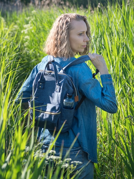 A view from the back of a blonde woman in a denim dress takes off his backpack on a path in the tall grass