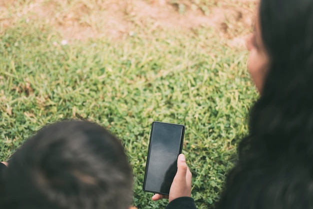 Photo view from the back group of laughing latinos sitting on the ground in a park with a smartphone