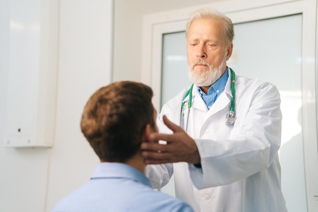 Vista dal retro al medico otorinolaringoiatra maschio adulto maturo toccando i linfonodi sul collo ed esaminando la gola del paziente di sesso maschile durante il controllo medico