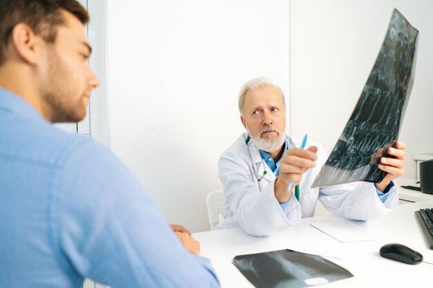 View from back of mature male doctor radiologist explaining results of MRI scanning for young man patient showing snapshot with images analyzing MRI scan in clinic physician reviewing test results