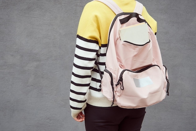 View from the back of an unrecognizable teenager girl with a school backpack against a gray wall
